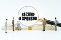 On a white surface, a dollar, figurines of people and a magnifying glass with the inscription -Become a Sponsor