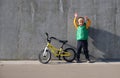 Ecstatic funny little child boy standing near concrete wall with his yelllow bicycle on sunny day. Urban lifestyle