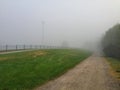 Eerie foggy silent early morning landscape with sandy footpath, green lawn, fence, bushes and streetlights Royalty Free Stock Photo