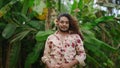 Flamboyant male with flower hairpiece strikes poses in floral shirt, denim shorts amidst lush greenery. Effeminate man