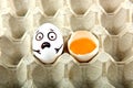 Egg with drawn cartoon faces and one broken egg in tray.