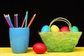 Eggs and pencils. Coloured crayons in blue mug and Easter decorations in green basket and red egg lying on yellow wooden Royalty Free Stock Photo