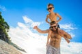 Eight years old boy sitting on dad`s shoulders. Both in swimming shorts and sunglasses, having fun on the beach. Bottom view