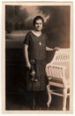 Vintage photo shows young woman poses next to historic carved bench. Black white antique studio photography.