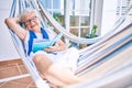 Elder senior woman with grey hair smiling happy relaxing on a hammock reading a book at home Royalty Free Stock Photo