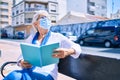 Elder senior woman with grey hair wearing coronavirus safety mask outdoors sitting on a bench reading a book Royalty Free Stock Photo