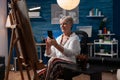 Elderly artist sitting down in front of easel with canvas browsing smartphone
