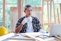 Elderly Asian carpenter with mustache taking a break and drinking a coffee, relaxing senior craftsman setting in working desk at