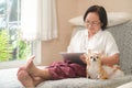 Elderly asian woman sitting on a sofa is using a tablet. She smiled happily, chihuahua dog sat on the side