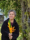 An elderly Asian woman smiling and holding yellow flowers while standing in a garden. Concept of aged people and relaxation Royalty Free Stock Photo