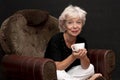 An elderly beautiful blonde woman of 60-65 years old holds a cup in her hand, sits in an armchair on a black background.