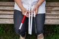 An elderly blind woman sits on a bench in the park. Close-up of female hands with folded tactile cane.