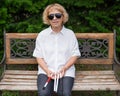 An elderly blind woman sits on a bench in the park with a folded tactile cane in her hands.