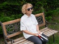 An elderly blind woman sits on a bench in the park with a folded tactile cane in her hands.