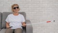 An elderly blind woman wearing sunglasses and with a tactile cane sits on the sofa.