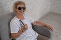 An elderly blind woman wearing sunglasses and with a tactile cane sits on the sofa.