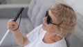 An elderly blind woman wearing sunglasses and with a tactile cane sits on the sofa.