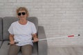 An elderly blind woman wearing sunglasses and with a tactile cane sits on the sofa.