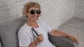 An elderly blind woman wearing sunglasses and with a tactile cane sits on the sofa.