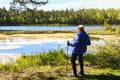 Elderly blond caucasian elderly woman stands holding red nordic sticks and enjoying lake forest green nature