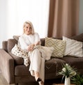elderly blond woman sit on couch at home holding knitting needles and yarn knits clothes for loved ones