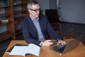 Elderly businessman grey hair in glasses sitting by the table, typing on laptop and holding mobile phone