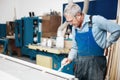 An elderly cabinetmaker in overalls and glasses paints a wooden board with a brush on a workbench in a carpentry shop.