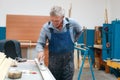 An elderly cabinetmaker in overalls and glasses paints a wooden board with a roller on a workbench in a carpentry shop.