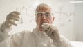 An elderly Caucasian male chemist in a protective suit holds a test tube and writes a formula on the glass. Royalty Free Stock Photo