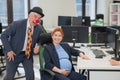 Elderly Caucasian man in clown costume amuses pregnant red-haired woman at work desk in office. Royalty Free Stock Photo