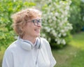 An elderly Caucasian woman walks in a park and listens to music. Smiling pensioner took off her headphones and enjoys