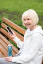 elderly cheerful woman using smartphone while sitting on bench with sport bottle