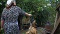 The elderly countrywoman plays with a puppy. The young dog sits on a chain. Sad landscape. Loneliness, poverty