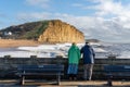 An elderly couple admire the view of the dramatic sunlit cliffs at West Bay, Dorset, UK.