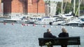 An elderly couple, a man and a woman with gray hair, sit on a bench by the lake on the background of yachts and modern houses.