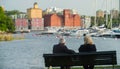 An elderly couple, a man and a woman with gray hair, sit on a bench by the lake on the background of yachts and modern houses.