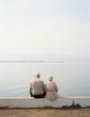 Elderly couple seated side by side on a brick wall, looking off into the distance, AI-generated.