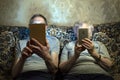 An elderly couple sit on the couch with gadgets, look at them close their faces.