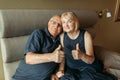 An elderly couple sits on a bed and shows a gesture with their hands - thumbs up, like