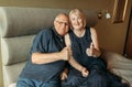 An elderly couple sits on a bed and shows a gesture with their hands - thumbs up, like