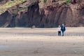 An elderly couple walk across an empty beach at the base of tall cliffs while holding hands