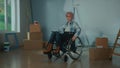 An elderly disabled woman in a wheelchair. Aged woman in room with window, cardboard boxes, wallpaper rolls, roller and