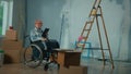 An elderly disabled woman in a wheelchair plans renovation using a digital tablet. Room with window, ladder, cardboard Royalty Free Stock Photo