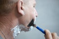 An elderly European man with foam on his face is shaving with a machine. Life of real people