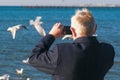 Elderly gray-haired man photographed on a mobile phone seascape with a pier and seagulls. Back view.