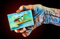 An elderly hand holds a painting made on a small canvas panel