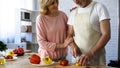 Elderly husband cutting fresh pepper in kitchen, wife hugging love, closeness