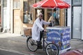 An elderly ice cream seller in a white robe rides a bicycle with his ice cream cart