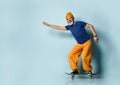 Elderly male in t-shirt, orange pants, hat, gumshoes. Riding black skateboard posing sideways on blue background. Full length