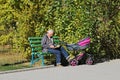 An elderly man with a baby in stroller sits in a park and reads a book